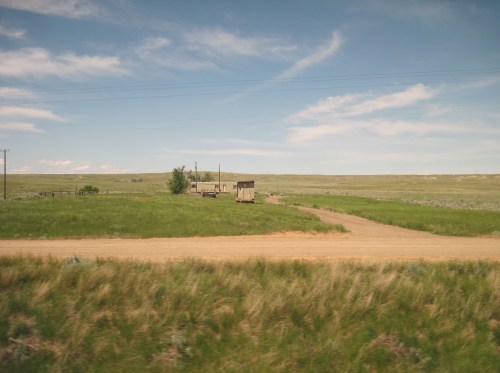 A dirt road winds through green fields, leading to a small, weathered building under a blue sky with scattered clouds.