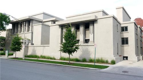 A modernist building with geometric shapes and textured concrete, featuring tall columns and greenery in front.