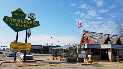 A retro-style hamburger restaurant with a large sign, featuring a cartoon duck, and an American flag in front.