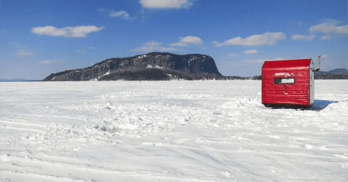 A red ice fishing hut on a snowy landscape with a distant mountain under a clear blue sky.