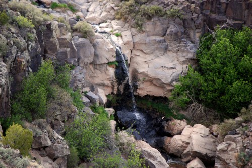 A small waterfall cascades down rocky cliffs into a lush green area surrounded by boulders and vegetation.