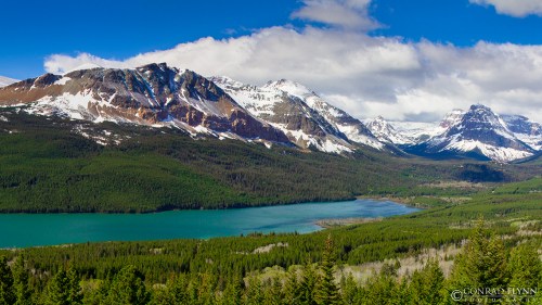 A panoramic view of snow-capped mountains surrounding a turquoise lake, with lush green forests in the foreground.