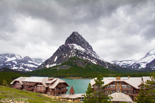 A scenic view of a mountain with snow-capped peaks, overlooking a lake and rustic buildings under a cloudy sky.