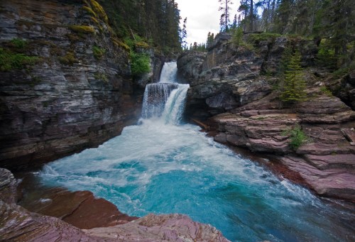 A serene waterfall cascades into a turquoise pool, surrounded by rocky cliffs and lush green trees.