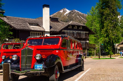 A vintage red bus parked in front of a lodge with a mountain backdrop under a clear blue sky.
