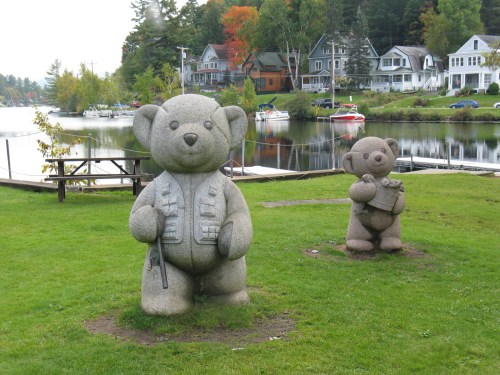 Two large teddy bear sculptures stand on a grassy area by a lake, with houses and boats in the background.