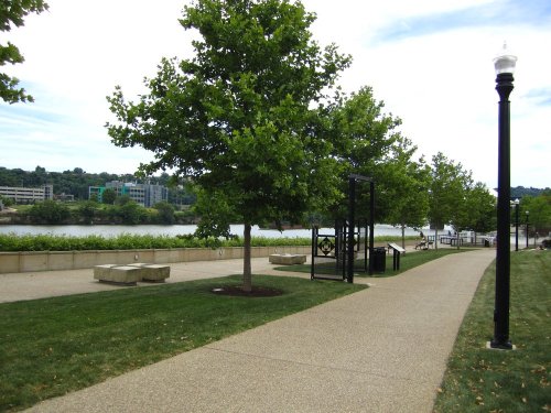 A scenic riverside path lined with trees, benches, and a view of the water under a partly cloudy sky.