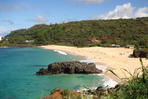 A scenic beach with golden sand, clear turquoise water, and lush green hills in the background under a blue sky.