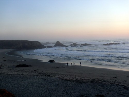 Three people walk along a sandy beach at sunset, with gentle waves and rocky formations in the background.