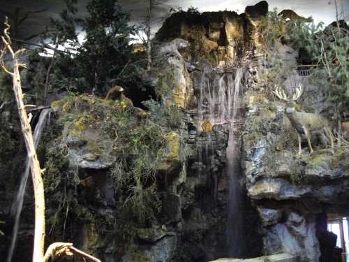 A rocky landscape with a waterfall, trees, and wildlife sculptures, creating a naturalistic scene indoors.