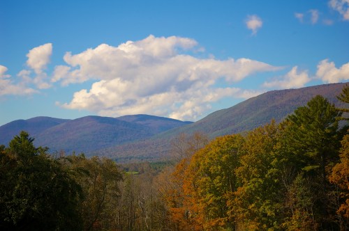 Scenic view of mountains under a blue sky with clouds, surrounded by autumn-colored trees.