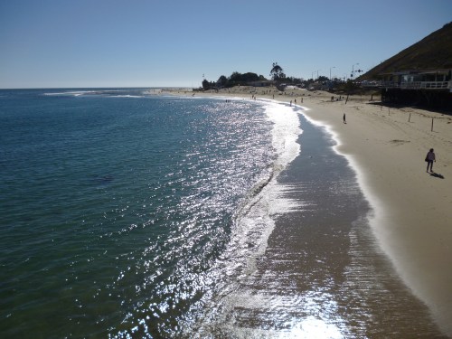 A sunny beach scene with gentle waves, a clear blue sky, and people walking along the shore.