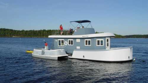 A modern houseboat with a blue exterior, docked on calm water, surrounded by trees and a clear sky.