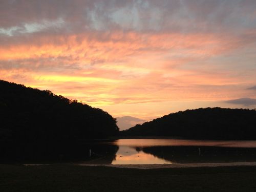 A serene lake at sunset, reflecting vibrant orange and pink hues against dark silhouettes of surrounding hills.