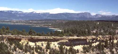 A panoramic view of a lake surrounded by mountains and lush green forests under a clear blue sky.