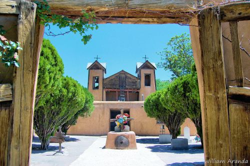 View through a wooden gate to a serene courtyard with a church, trees, and a colorful flower arrangement.