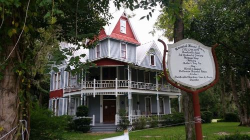 A historic Victorian-style house with a red roof, surrounded by greenery and a sign detailing its history.
