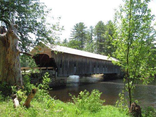 A wooden covered bridge spans a river, surrounded by lush greenery and tall trees under a clear blue sky.