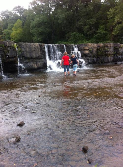 Two children play in shallow water near a small waterfall, surrounded by lush greenery and rocky terrain.