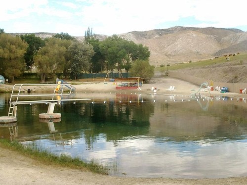 A serene swimming area with diving boards, surrounded by trees and mountains, featuring a sandy beach and calm water.