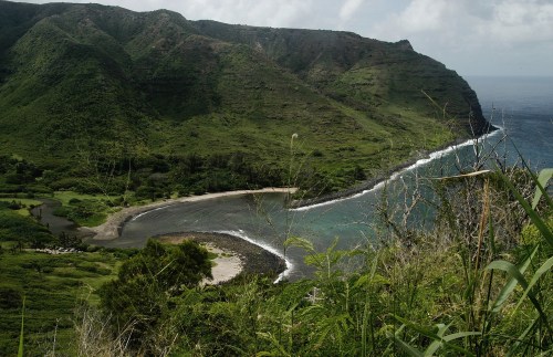Lush green hillsides meet a tranquil bay, with gentle waves lapping at the shore under a cloudy sky.