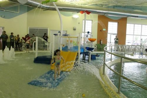 A child runs through a splash pad area with water features and colorful play structures in a recreational facility.