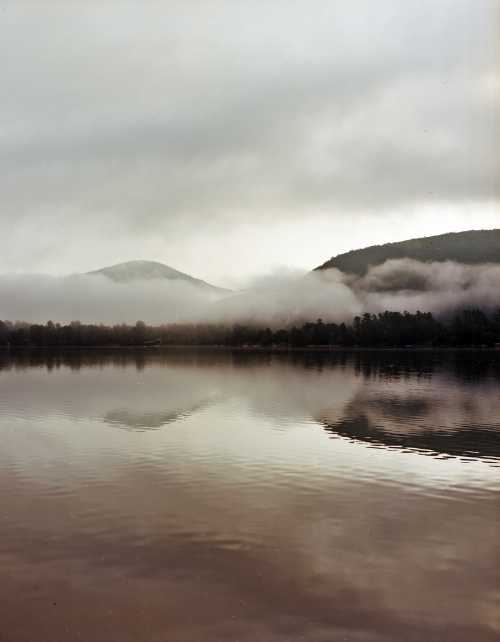 A serene lake reflects misty mountains and clouds under a gray sky, creating a tranquil, atmospheric scene.