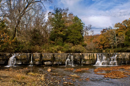 A serene landscape featuring a waterfall cascading over rocks, surrounded by autumn foliage and a clear blue sky.