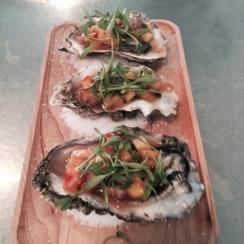 Three oysters on a wooden platter, topped with mango salsa and microgreens, garnished with salt.