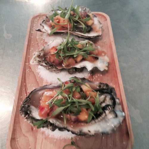 Three oysters on a wooden platter, topped with mango salsa and microgreens, garnished with salt.