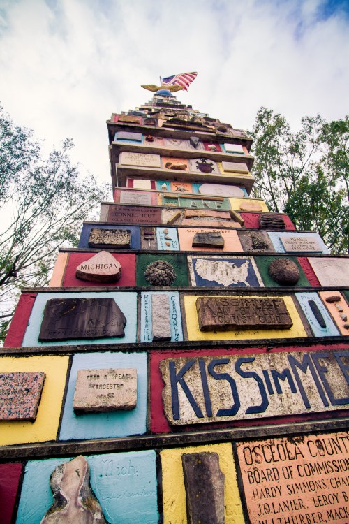 A colorful monument with various plaques and an American flag at the top, surrounded by trees and a blue sky.