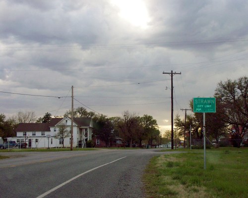 A rural road leads to a city limit sign for Strawn, with houses and trees under a cloudy sky.