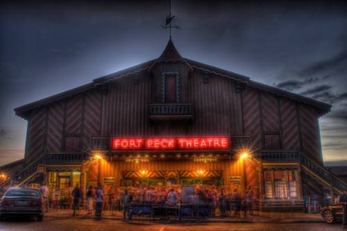 The Fort Peck Theatre illuminated at dusk, with a crowd gathered outside and a vibrant marquee sign.