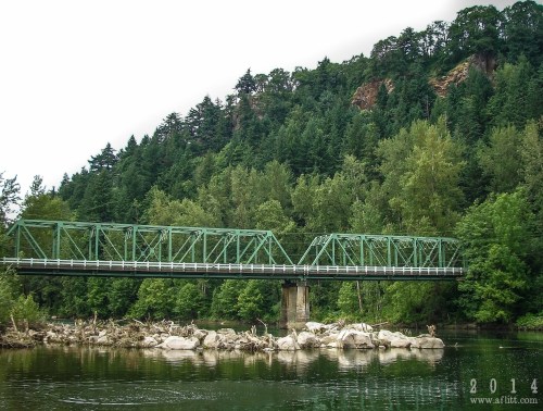 A green metal bridge spans a calm river, surrounded by lush trees and rocky banks under a cloudy sky.