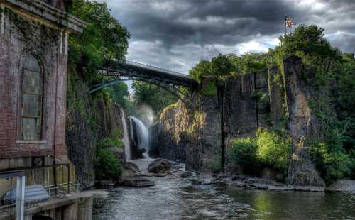 A scenic view of a waterfall surrounded by rocky cliffs and greenery, with a bridge and an American flag in the background.