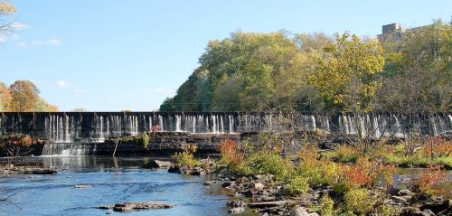 A serene waterfall cascades over a dam, surrounded by colorful autumn foliage and a calm river.