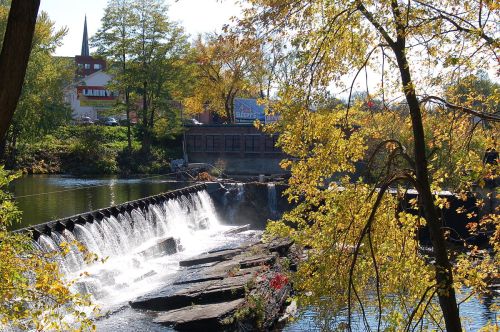 A serene river scene featuring a small waterfall, surrounded by autumn foliage and buildings in the background.