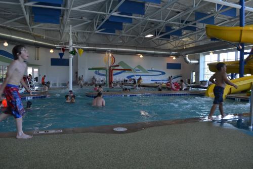 Indoor water park scene with children swimming, playing, and a yellow water slide in the background.