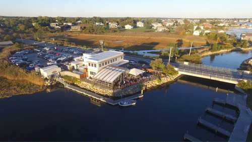 Aerial view of a waterfront restaurant with outdoor seating, surrounded by water and parked cars nearby.