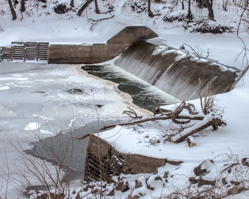 A frozen river with a concrete dam, surrounded by snow-covered banks and scattered branches.