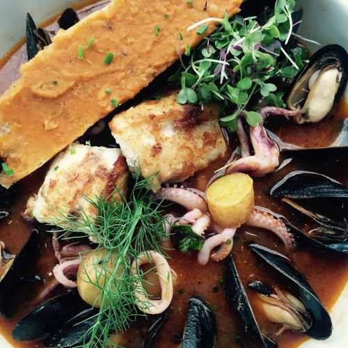 A bowl of seafood stew featuring fish, mussels, squid, and garnished with herbs and a crispy topping.