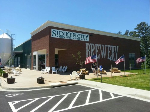 Exterior of Sunken City Brewing Company with American flags, picnic tables, and a clear blue sky.