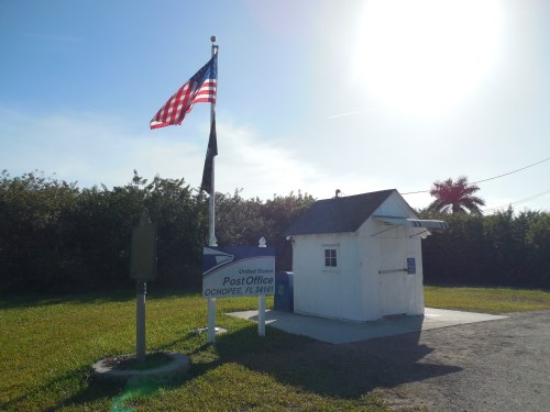 A small white post office building with an American flag and a sign in a grassy area surrounded by trees.