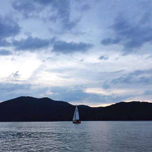 A sailboat glides across calm waters under a cloudy sky, with mountains in the background.