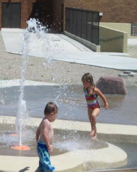 Two children play in a splash pad, with water spraying around them on a sunny day.