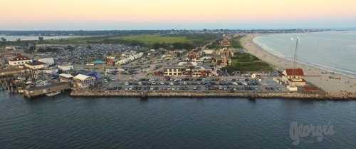 Aerial view of a beachside area with a pier, parking lot, and coastline under a pastel sunset sky.