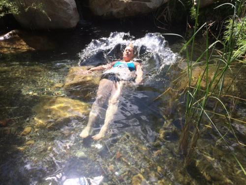 A person relaxes in a clear stream, surrounded by rocks and greenery, with water splashing around them.