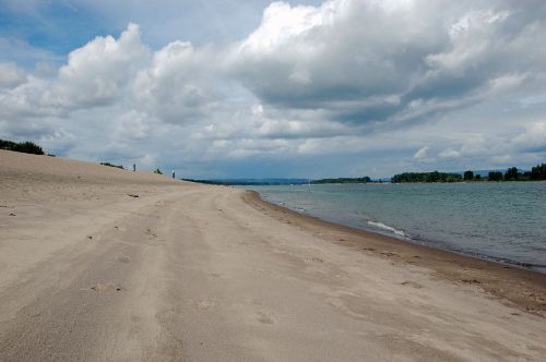 A sandy beach curves along a riverbank under a cloudy sky, with distant trees lining the horizon.