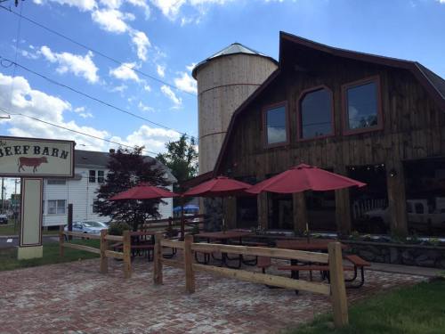 A rustic restaurant with a wooden exterior, outdoor seating under red umbrellas, and a silo in the background.