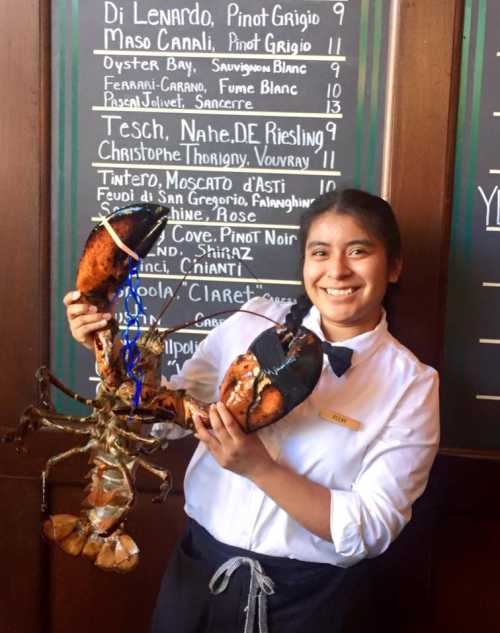 A smiling server in a white shirt holds a large lobster in front of a chalkboard menu.